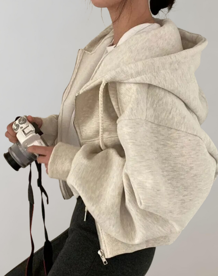 Světle šedá melírovaná mikina na zip z boku, model drží fotoaparát, vidět objemné rukávy a kapuci. 2