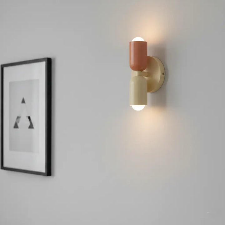 Detail rozsvíceného lososovo-krémového svítidla na šedé stěně vedle černě zarámovaného obrazu s minimalistickým motivem. 4