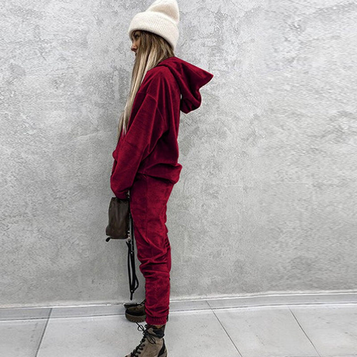 Frau in rotem Hoodie und Jogginghose, weiße Mütze, braune Stiefel, grauer Hintergrund.