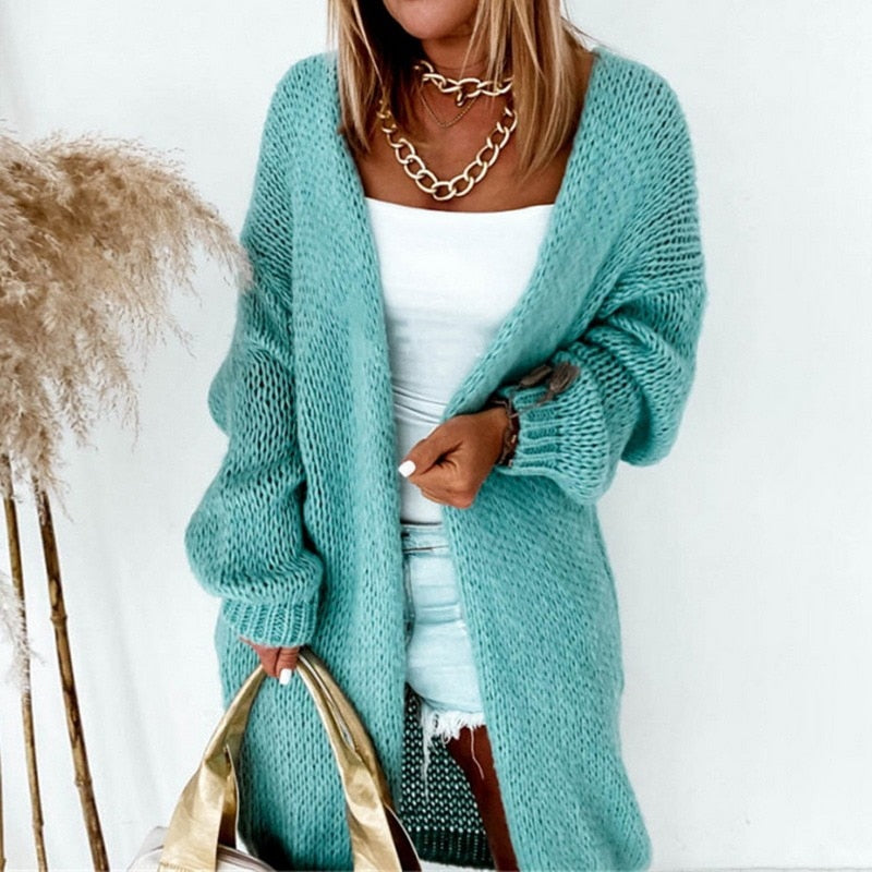 Frau in türkisfarbenem Strickcardigan, weißem Top und Jeansshorts, hält goldene Tasche.