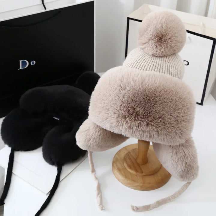 Beige Wintermütze mit Bommel, Kunstfell, warm, stilvoll, auf Holzständer, neben schwarzer Mütze.