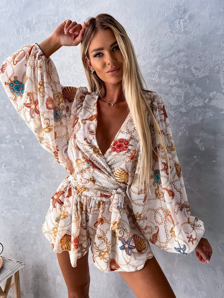 Frau in weißem, gemustertem Sommerkleid mit Muschel- und Kettenmotiv vor grauer Wand.