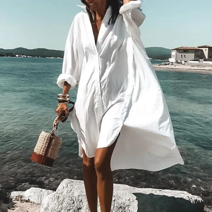 Frau in weißem Sommerkleid am Strand, trägt Korbtasche, Meer im Hintergrund.