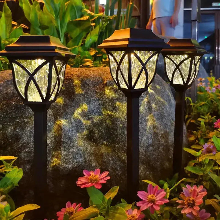 Drei schwarze Solar-Gartenleuchten bei Nacht, umgeben von Blumen und Pflanzen.
