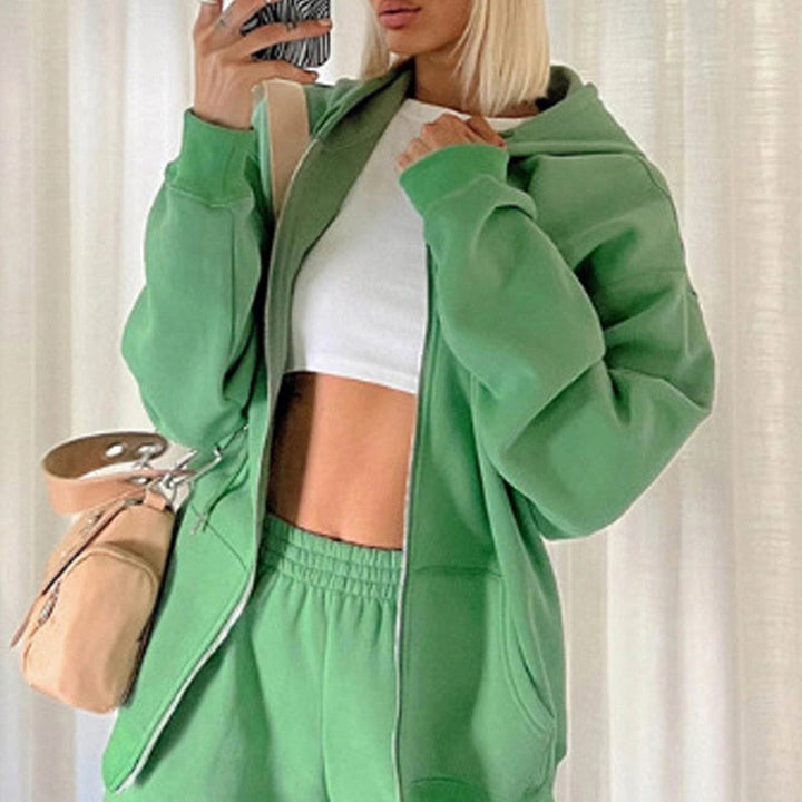 Frau in grünem Jogginganzug mit Kapuze, weißem Top, beige Tasche, Selfie im Spiegel.