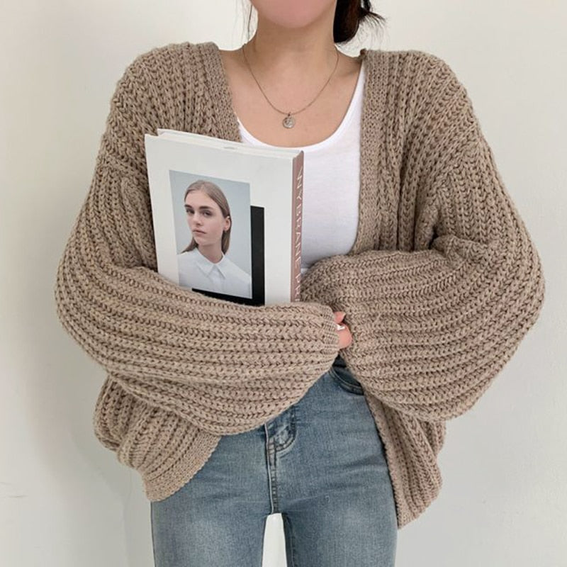 Frau in beigem Strickcardigan, hält Buch, trägt weißes Shirt und Jeans.