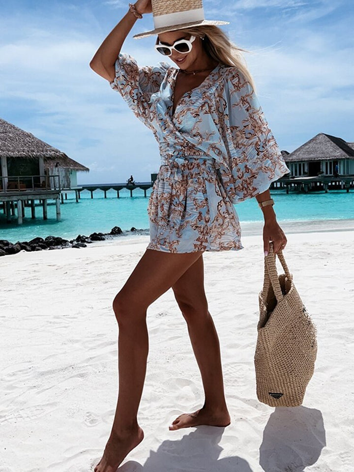 Frau im blauen Strandkleid mit Strohhut und Tasche am tropischen Strand vor Wasserbungalows.