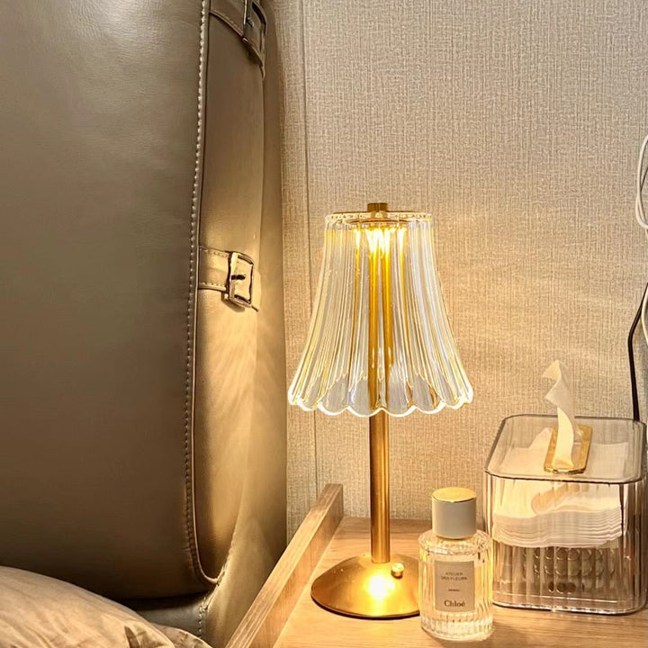 Goldene Tischlampe mit Glaslampenschirm auf Nachttisch, neben Parfümflasche und Taschentuchbox.