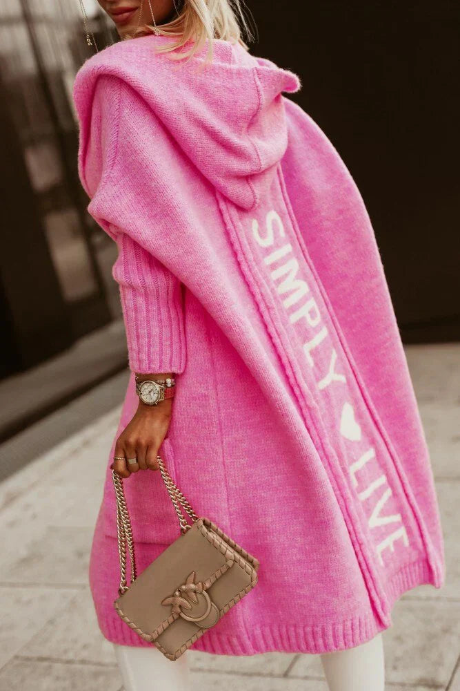 Frau in pinkem Strickmantel mit Kapuze und "SIMPLY LIVE"-Schriftzug, beige Handtasche.