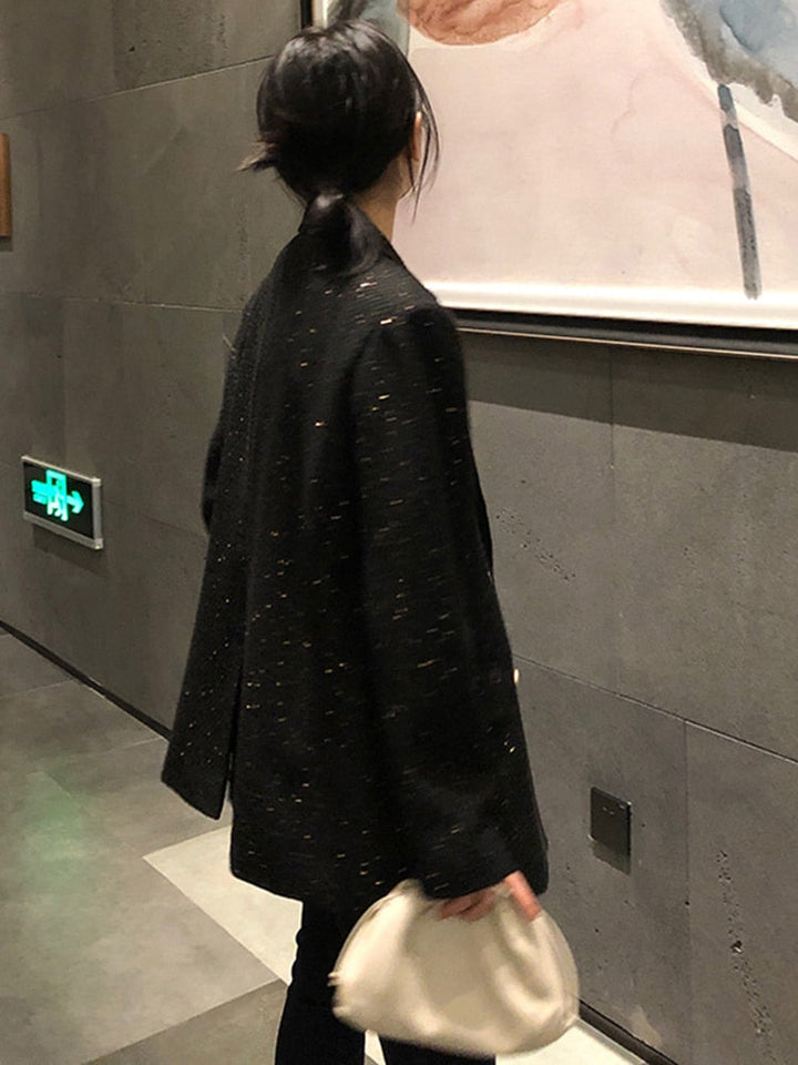 Frau in schwarzem Blazer mit weißer Tasche betrachtet Kunstwerk an grauer Wand.