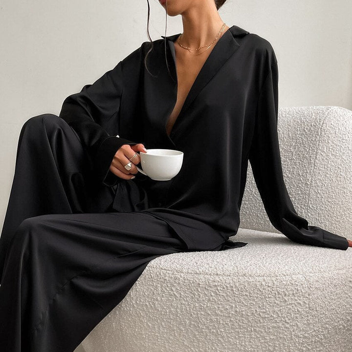 Frau in schwarzem Satin-Pyjama hält weiße Tasse, sitzt auf modernem Sessel.
