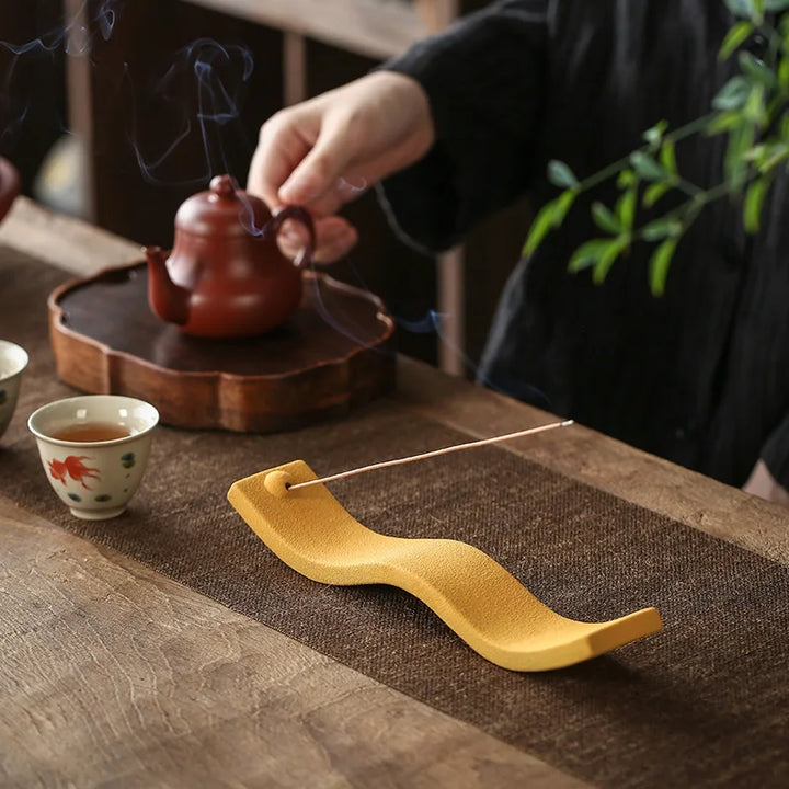 Gelber Keramik-Räucherstäbchenhalter auf Holztisch, neben Teekanne und Tasse, Zen-Dekor.