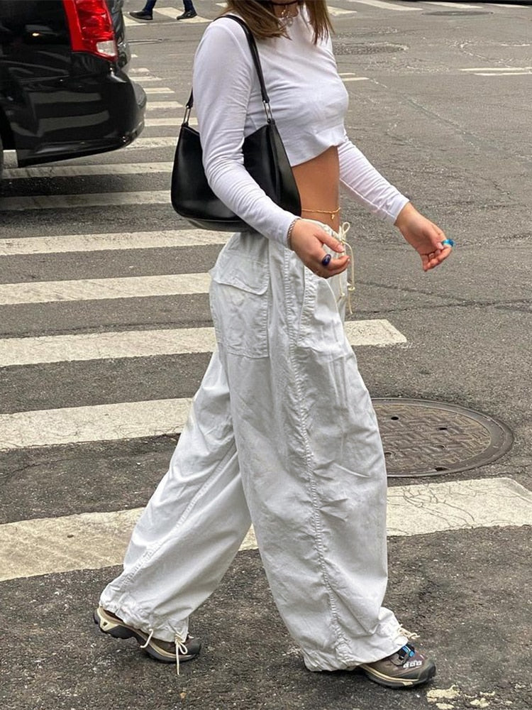Frau in weißem Crop-Top und weiten weißen Hosen, schwarze Tasche, Sneakers, Straßenhintergrund.