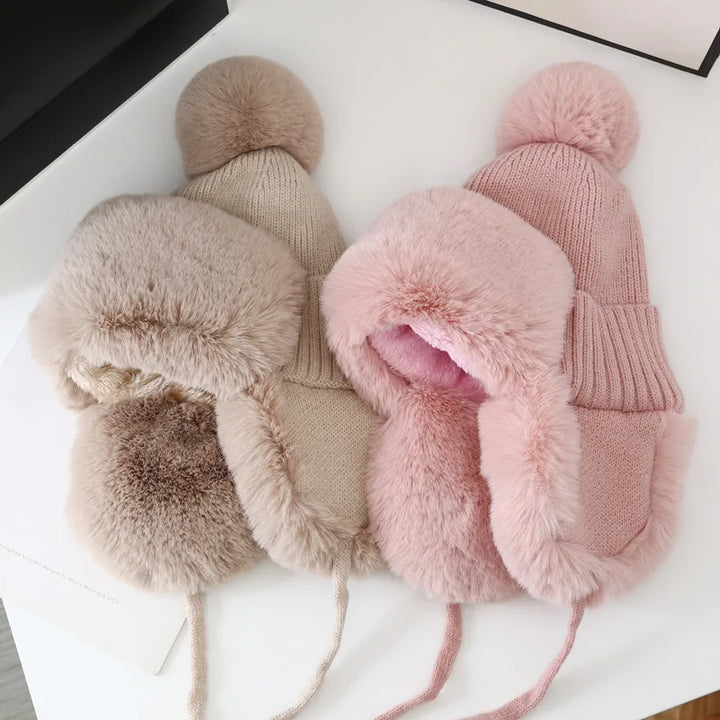 Zwei Wintermützen aus Strick und Kunstfell in Beige und Rosa mit Bommel auf weißem Hintergrund.