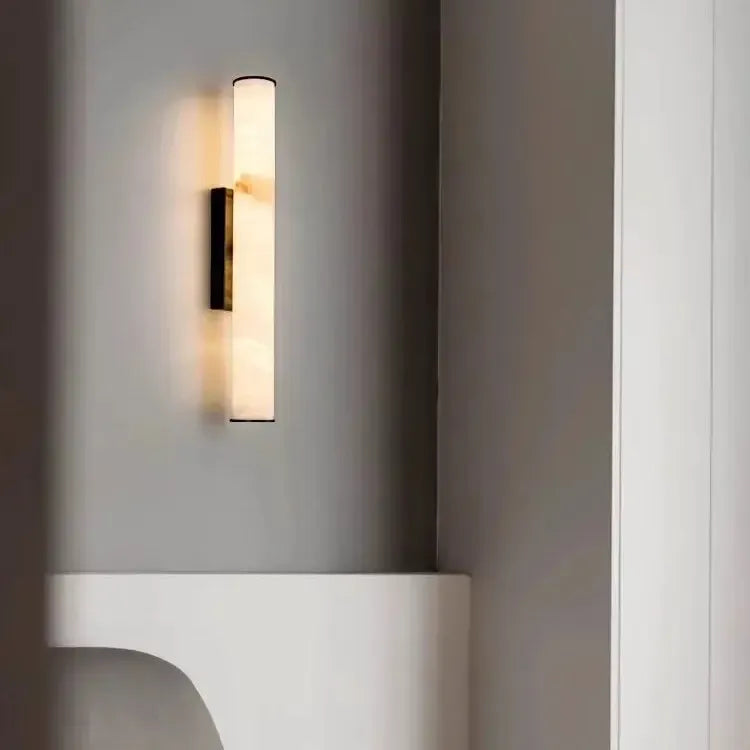 Svítidlo s tmavým držákem na šedé stěně nad bílým zaobleným nábytkem, vedle světlého sloupku. 20
