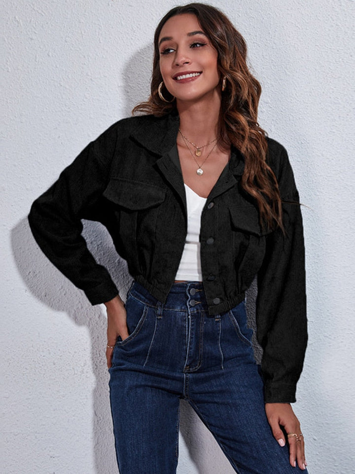 Frau in schwarzer Jeansjacke, weißem Top und blauer Jeans vor weißer Wand.