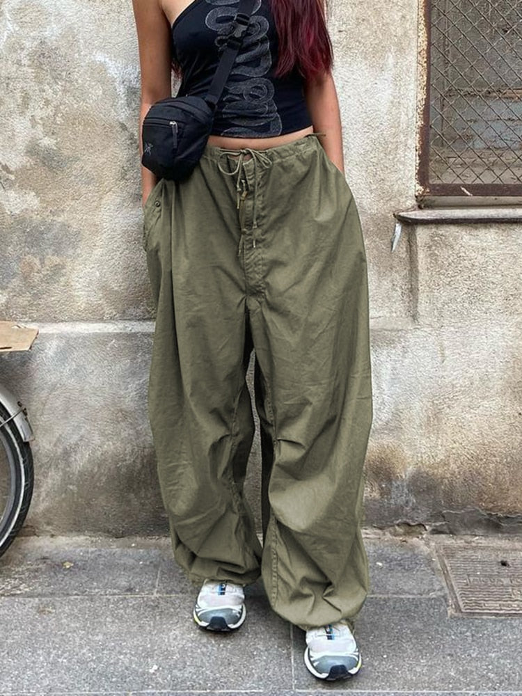 Frau in olivgrüner Baggy-Hose, schwarzem Top, Sneaker und Bauchtasche vor Betonwand.