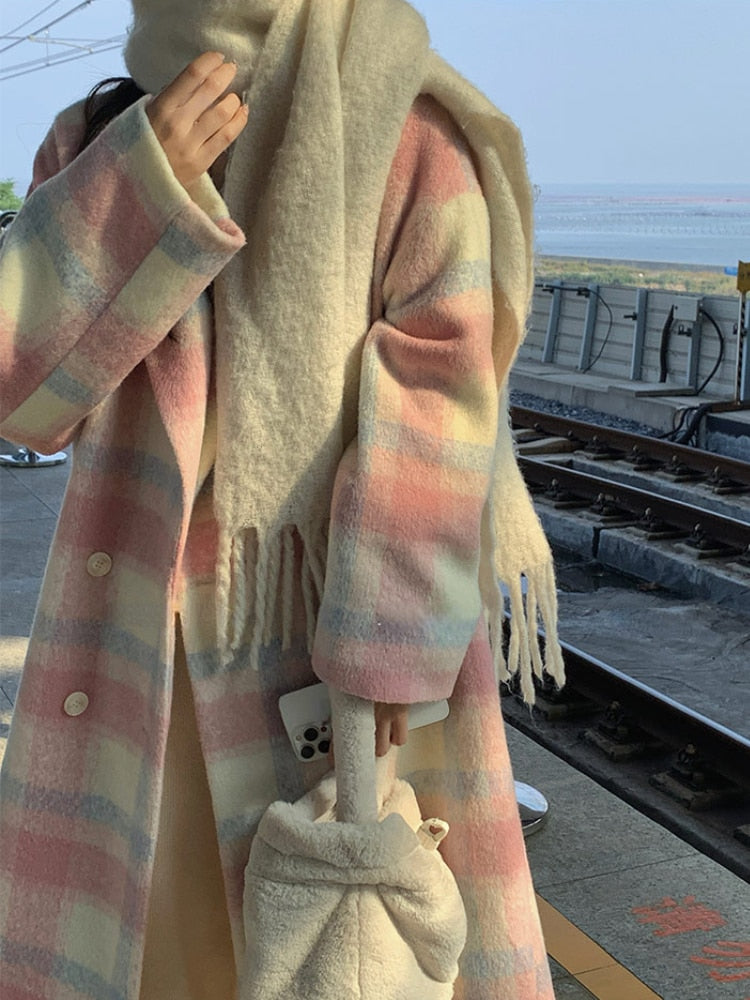 Frau im pastellfarbenen karierten Mantel mit weißem Schal und Tasche am Bahnhof.
