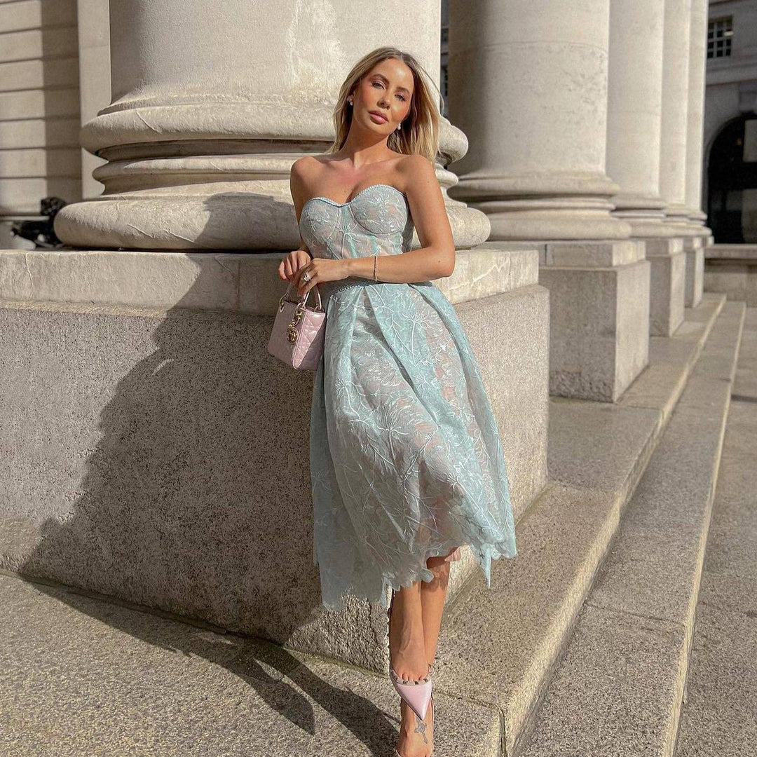 Frau in hellblauem Spitzenkleid vor Säulen, hält rosa Handtasche, elegantes Sommeroutfit.