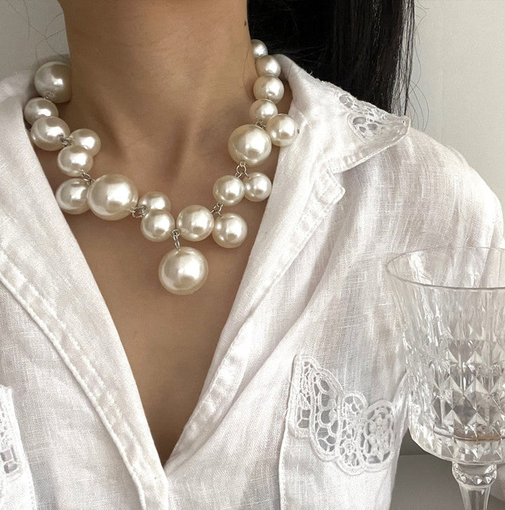 Frau trägt weiße Perlenkette, elegantes Design, auf weißer Bluse, hält Kristallglas.