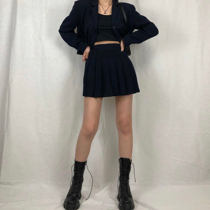 Frau in schwarzem Blazer und Faltenrock, schwarze Stiefel, modisches Outfit, weißer Hintergrund.