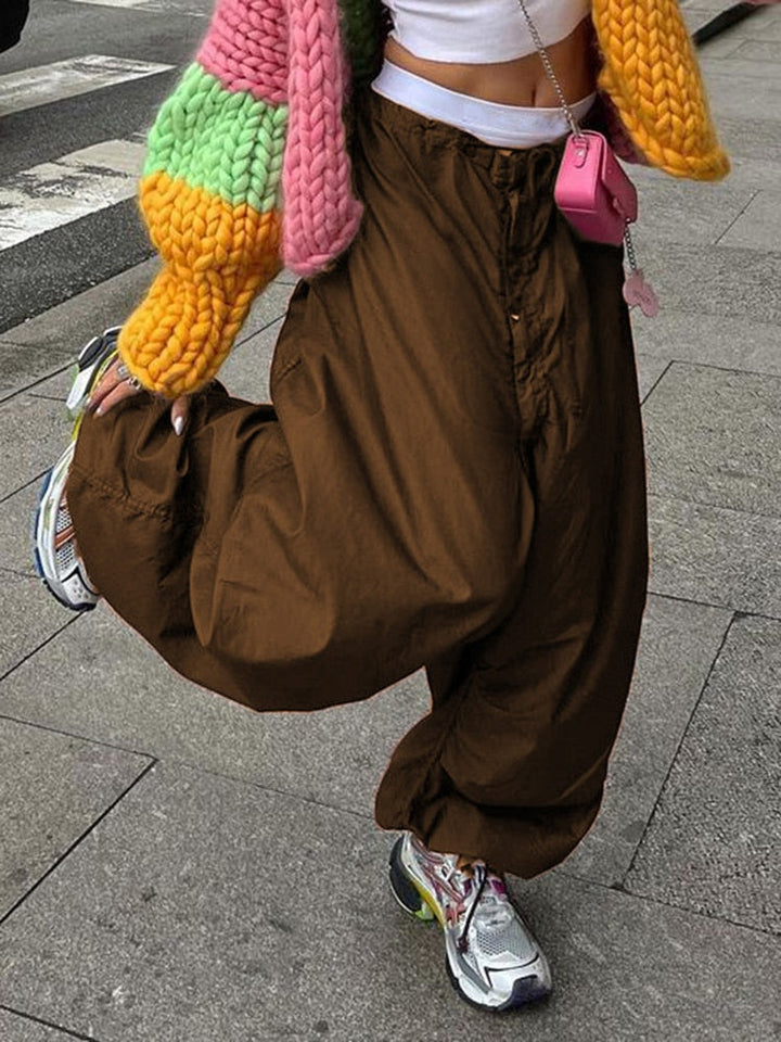 Frau in brauner Baggy-Hose, bunter Strickjacke, weiße Sneakers, pinke Umhängetasche auf Gehweg.