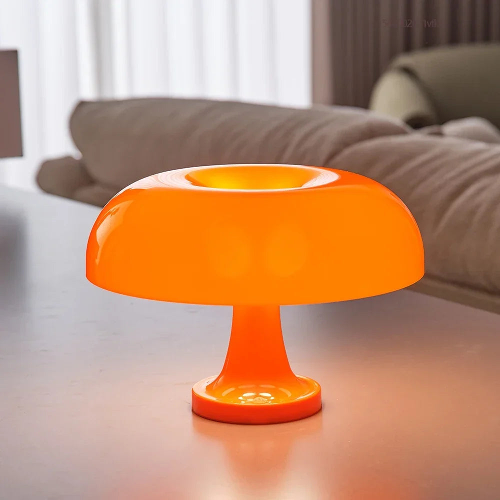 Oranžová rozsvícená lampa s širokým lesklým stínidlem a prohlubní nahoře stojí na stole před béžovou pohovkou. 3