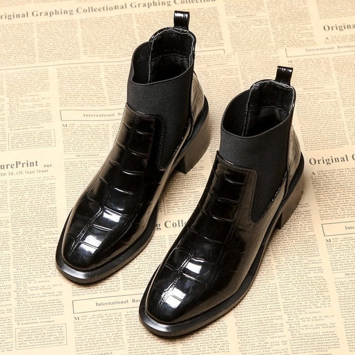 Schwarze Chelsea-Stiefel aus Leder mit Krokodilprägung auf Zeitungshintergrund.