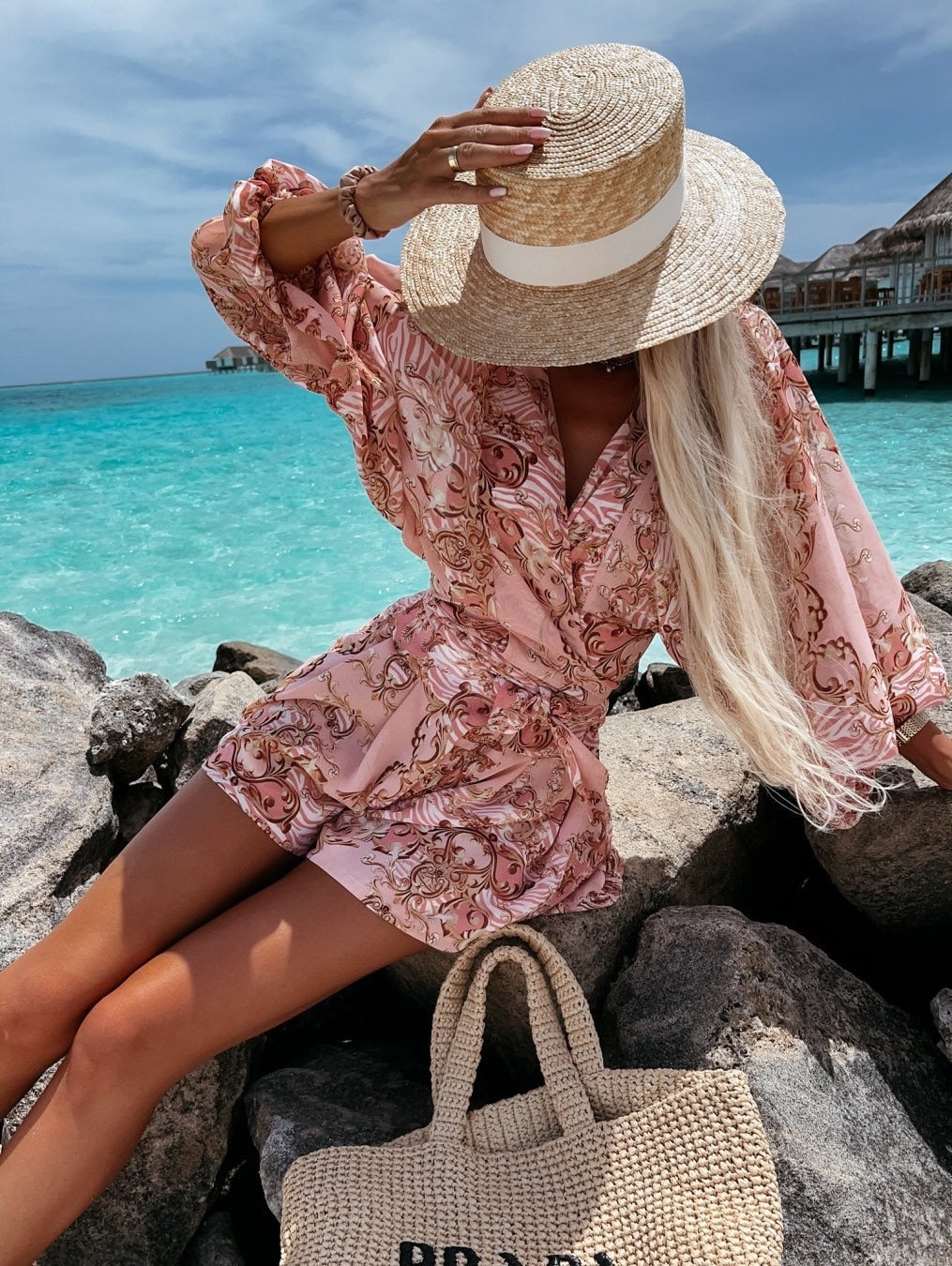 Frau in rosa Sommerkleid und Strohhut am Strand, türkisfarbenes Meer im Hintergrund.