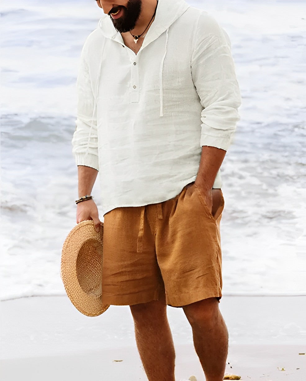 Mann am Strand in weißem Leinenhemd und braunen Shorts, hält Strohhut.