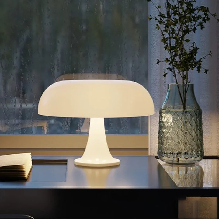 Bílá rozsvícená houbová lampa stojí na tmavém stole vedle otevřené knihy a skleněné vázy s větvičkami, za oknem prší. 4