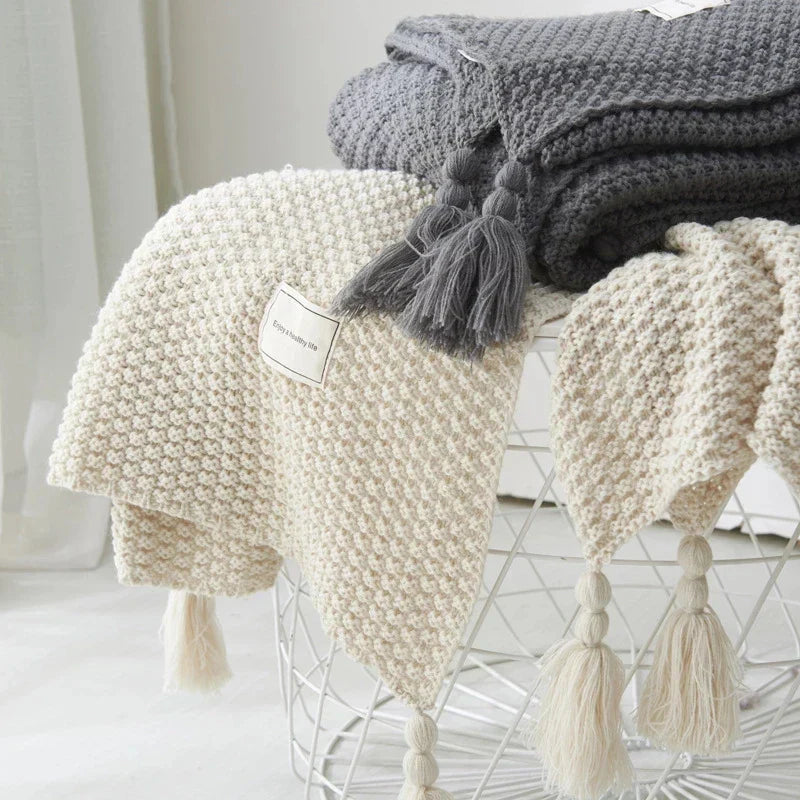 Beige und graue Strickdecken mit Quasten auf weißem Drahtkorb, gemütliche Wohnaccessoires.
