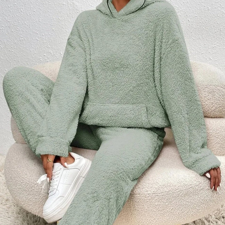 Frau in grünem Fleece-Hoodie und Jogginghose auf Sofa, weiße Sneaker, gemütliche Mode.