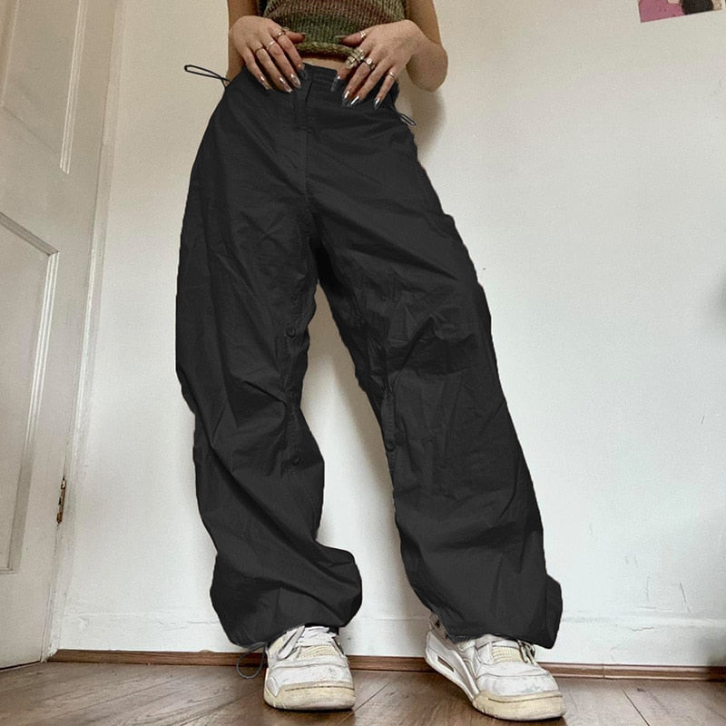 Frau trägt schwarze Baggy-Hose, weiße Sneaker, lässiger Streetwear-Look, Holzboden.
