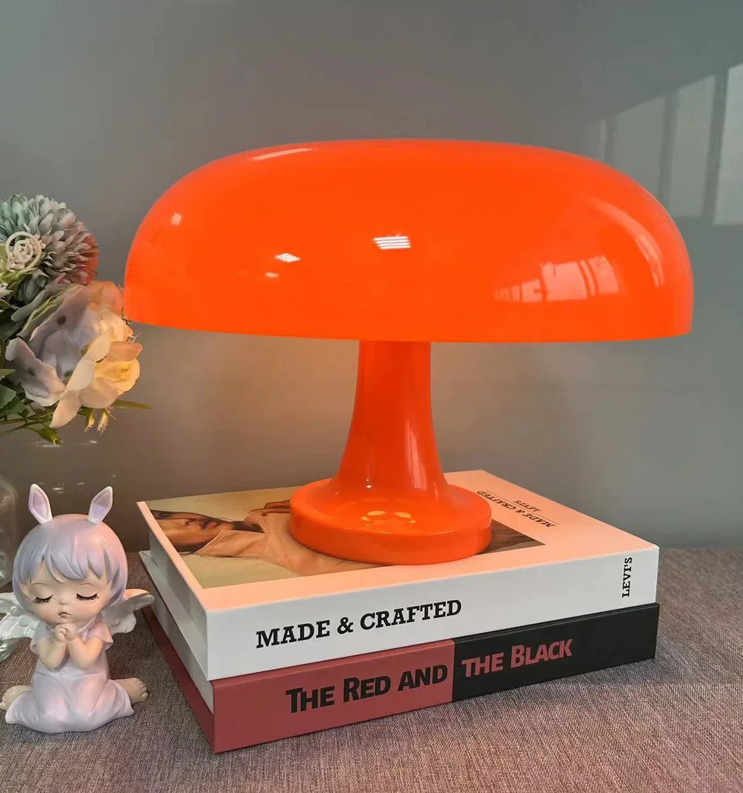 Oranžová lampa stojí na dvou knihách vedle kytice a figurky andílka s králičíma ušima. 7