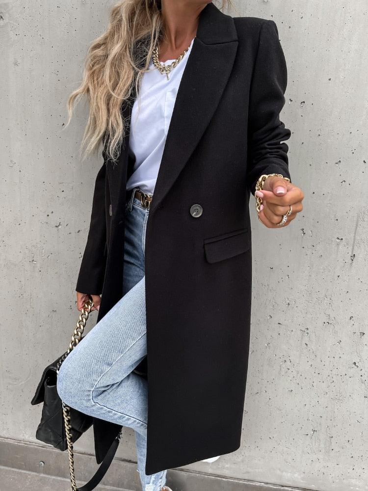 Frau in schwarzem Wollmantel, weiße Bluse, Jeans, goldene Accessoires, modisch, elegant.