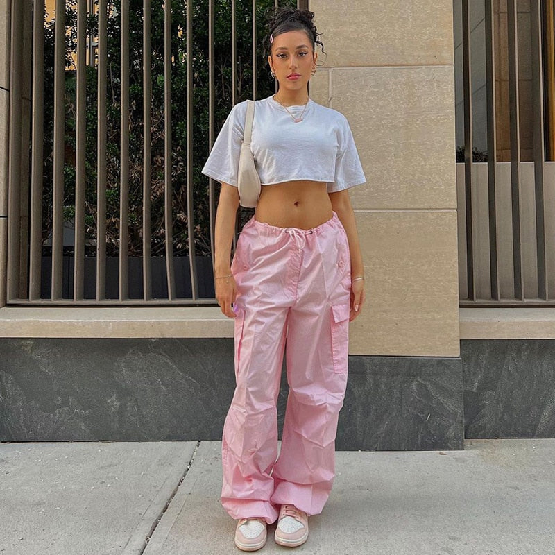 Frau in weißem Crop-Top und rosa Cargo-Hose, lässiger Streetwear-Look, urbaner Hintergrund.