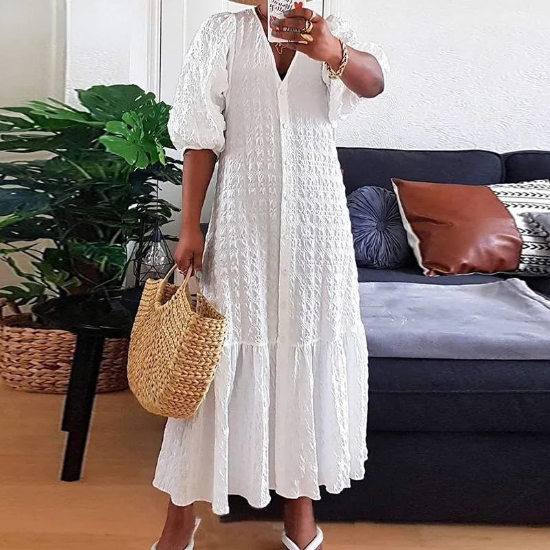 Frau in weißem Sommerkleid mit Strohtasche vor Sofa und Zimmerpflanze.