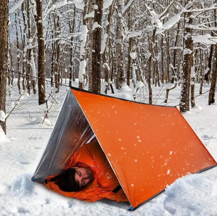 Mann in orangefarbenem Notfallzelt im verschneiten Wald, Wintercamping, Überlebensausrüstung.