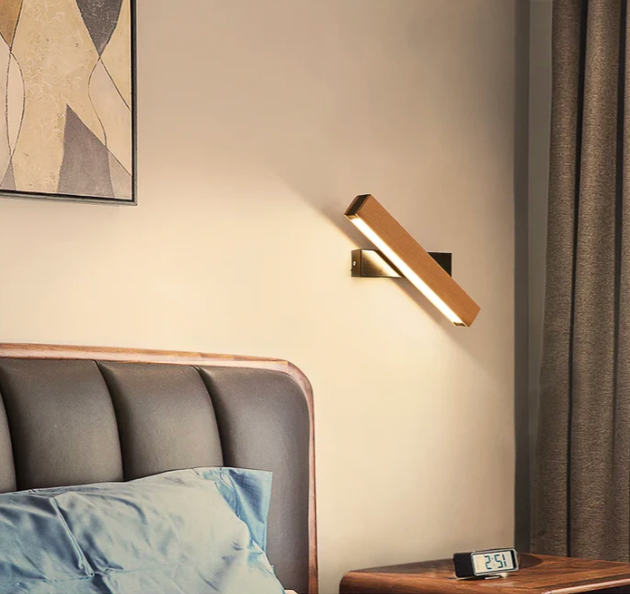 Nakloněná lampa s tmavým držákem svítí nad čalouněným čelem postele, vedle noční stolek s digitálními hodinami. 8