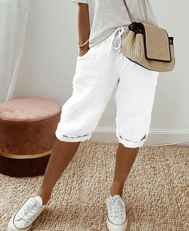 Frau in weißer Leinenhose, grauem T-Shirt, Strohtasche, weiße Sneaker, lässiger Sommerlook.