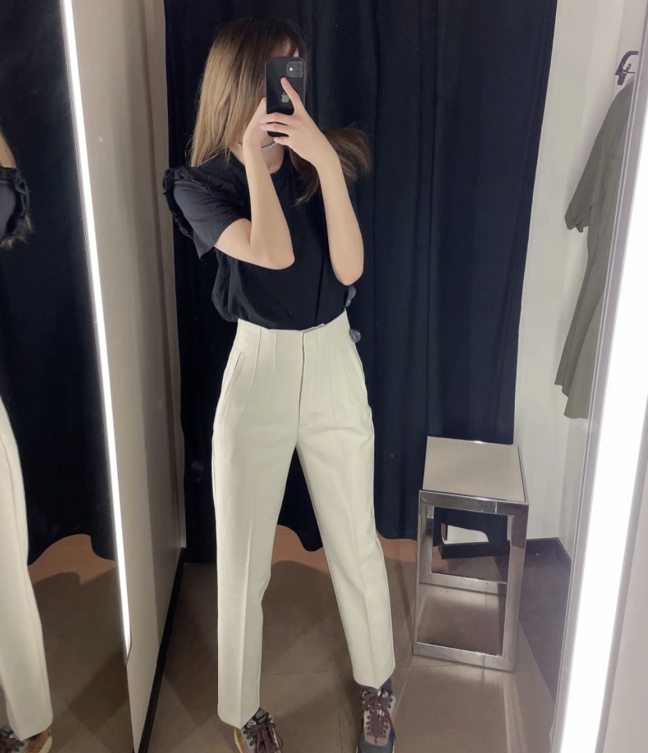 Frau in Umkleidekabine mit schwarzem Oberteil und weißen Hosen, Spiegel-Selfie, Mode.
