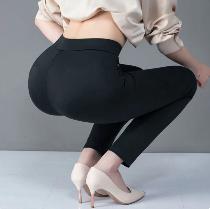 Frau in schwarzer Leggings und beigen High Heels, kniend auf grauem Hintergrund.