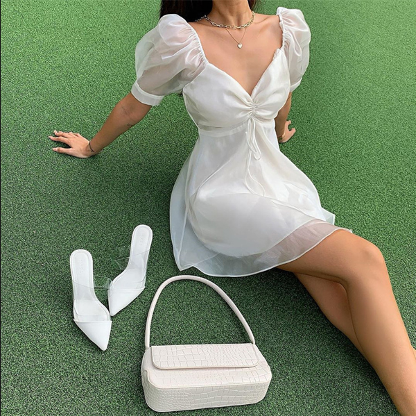 Frau in weißem Sommerkleid mit Puffärmeln, weiße Handtasche und Schuhe auf grünem Rasen.