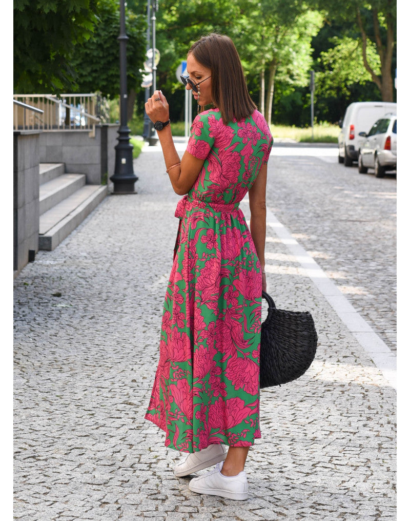 Frau im pink-grünen Blumenkleid, trägt schwarze Tasche, weiße Sneaker, steht auf gepflasterter Straße.