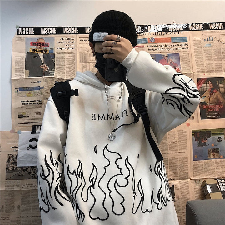 Person in weißem Hoodie mit Flammenmuster, schwarzer Mütze, Smartphone, vor Zeitungswand.
