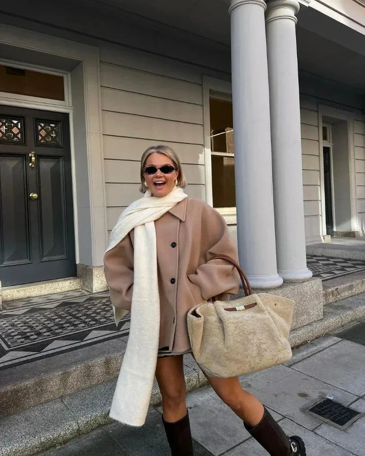 Frau in beiger Jacke mit weißem Schal und großer Tasche vor Gebäude mit Säulen.