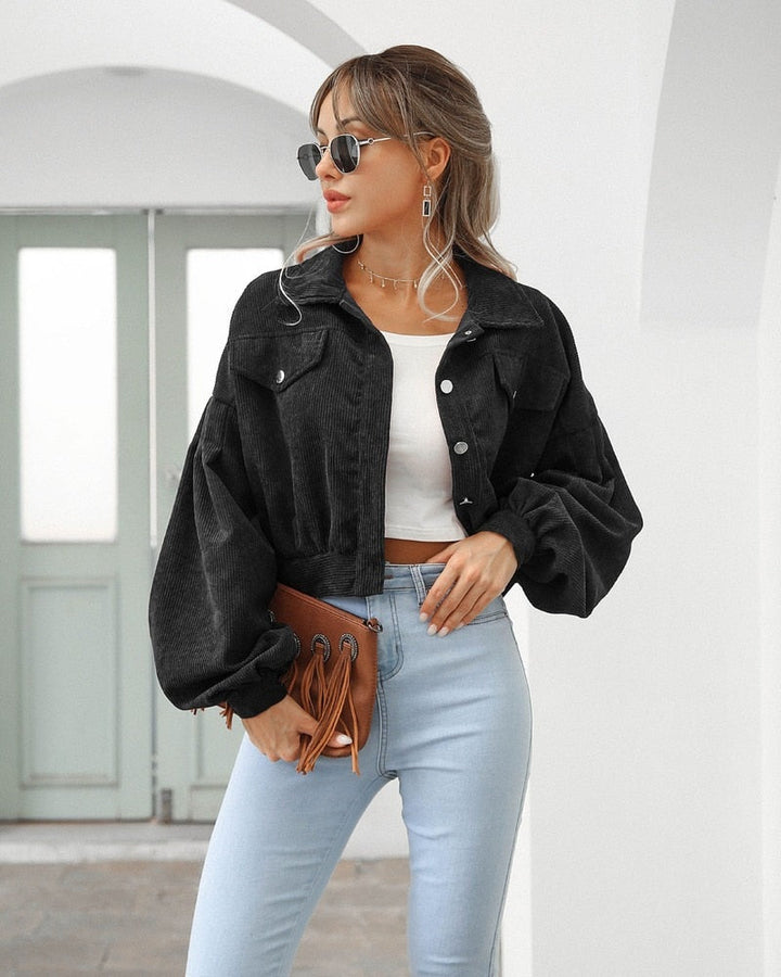 Frau in schwarzer Cordjacke, weißem Top, blauer Jeans, Sonnenbrille, braune Tasche, modisch.