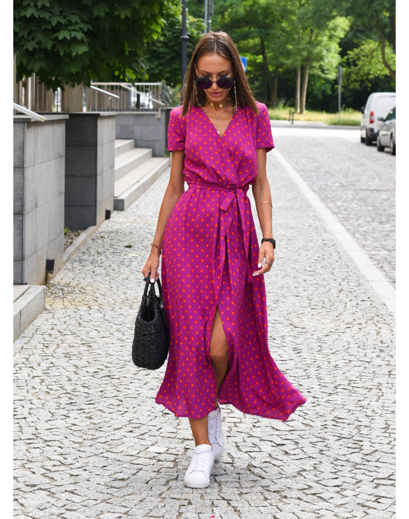 Frau in pinkem Wickelkleid mit Punkten, trägt schwarze Tasche, weiße Sneaker, Sonnenbrille.