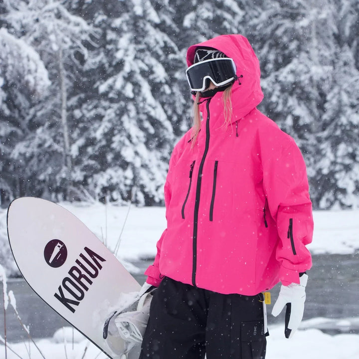 Modelka v neonově růžové bundě s kapucí drží snowboard, v pozadí zasněžené stromy. 5